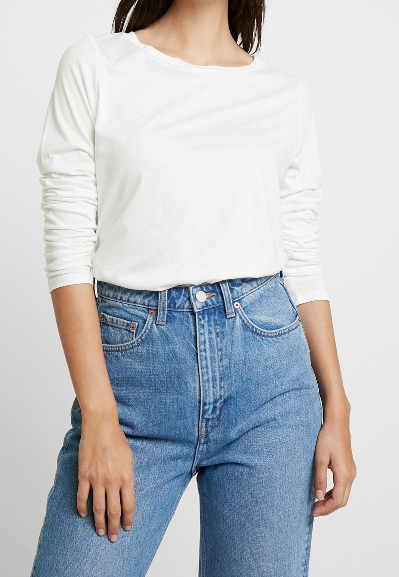 Langärmliges weißes Baumwollhemd mit Rundhalsausschnitt, kombiniert mit hochgeschnittenen blauen Jeans aus Denim, die klassische Taschen und einen verwaschenen Look aufweisen.