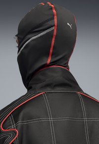Tuta da corsa nera con cappuccio fitted e dettagli rossi. Presenta cuciture bianche a contrasto e il logo Puma sul retro. Tessuto testurizzato visibile.