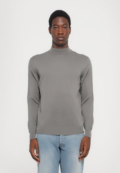 Maglione grigio lavorato a maglia con un collo alto, maniche lunghe e una vestibilità aderente, abbinato a jeans azzurri chiari. Texture morbida e dettagli minimi.