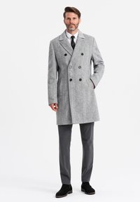 Abrigo de lana gris de mezcla, de doble botonadura, con solapas de muesca, botones negros, bolsillos laterales y un corte entallado, combinado con pantalones grises y botas negras.