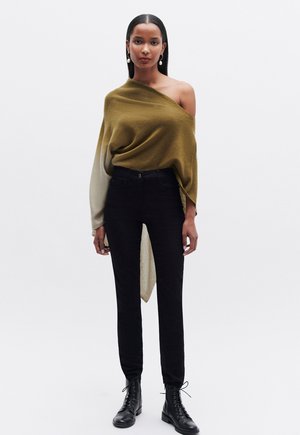 Olijfgroene en beige gedrapeerde trui met één schouder bloot, gecombineerd met strakke zwarte jeans en zwarte veterschoenen. Glad oppervlak en casual ontwerp.