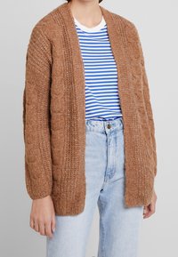 Brown cable-knit cardigan with a loose fit, open front, and ribbed accents. Worn over a blue and white striped shirt and light jeans.