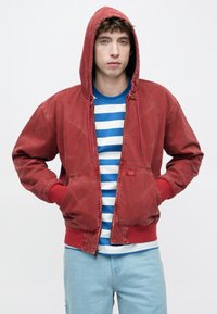 Κόκκινο μπουφάν με κουκούλα και κορδόνια, μπροστινές τσέπες, ελαφρώς φθαρμένο, συνδυασμένο με μπλε και λευκή ριγέ μπλούζα, ανοιχτόχρωμο τζιν παντελόνι.