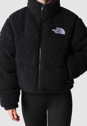 Persona que lleva una chaqueta negra esponjosa con cremallera y el logo de The North Face en el pecho izquierdo, combinada con pantalones negros sobre un fondo liso.