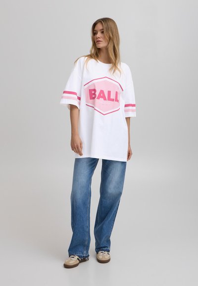 Camiseta blanca de gran tamaño con el logo rosa "BALL", con mangas cortas que tienen rayas rosas. Combinada con jeans de pierna ancha azules y zapatos beige.