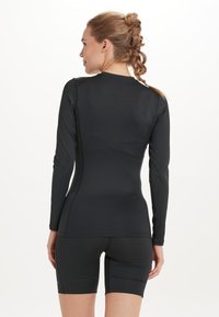 Langärmliges, schwarzes Sportoberteil mit figurbetontem Schnitt, kombiniert mit schwarzen Shorts. Das Material wirkt glatt, dehnbar und nahtlos.