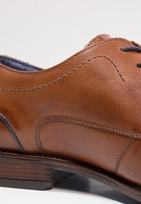 Brauner Lederschuh mit glatter Textur, abgerundetem Zeh, dekorativen Nähten und blauen Akzenten an der Seite. Absatz aus geschichtetem Holz.