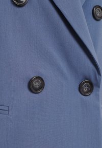 Tessuto del blazer azzurro chiaro con una texture liscia, caratterizzato da due grandi bottoni neri e una tasca laterale. Design su misura e strutturato.