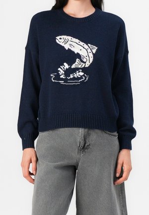 Pull bleu marine avec un graphisme blanc d'un poisson sautant hors de l'eau, porté par une personne en pantalon gris, faisant face vers l'avant.