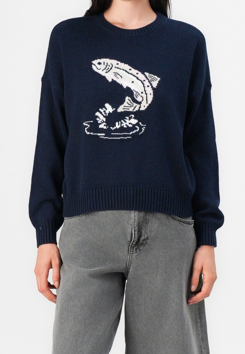 Pull bleu marine avec un graphisme blanc d'un poisson sautant hors de l'eau, porté par une personne en pantalon gris, faisant face vers l'avant.