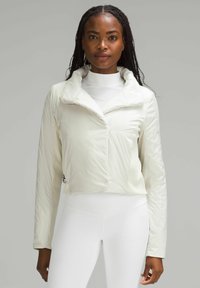 Krótkie białe puchowe kurtki z wysokim kołnierzem, gładką teksturą i prążkowanymi akcentami na rękawach, noszonej na białym golfie i legginsach.