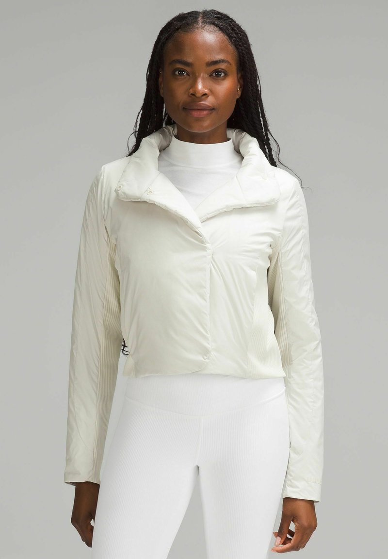 Krótkie białe puchowe kurtki z wysokim kołnierzem, gładką teksturą i prążkowanymi akcentami na rękawach, noszonej na białym golfie i legginsach.