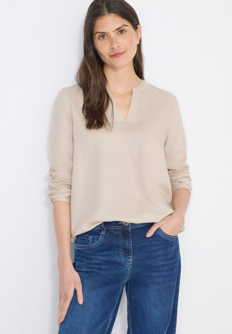 Blouse beige à manches longues avec un col en V, une texture lisse et des manches retroussées, associée à un jean denim bleu avec des poches avant.
