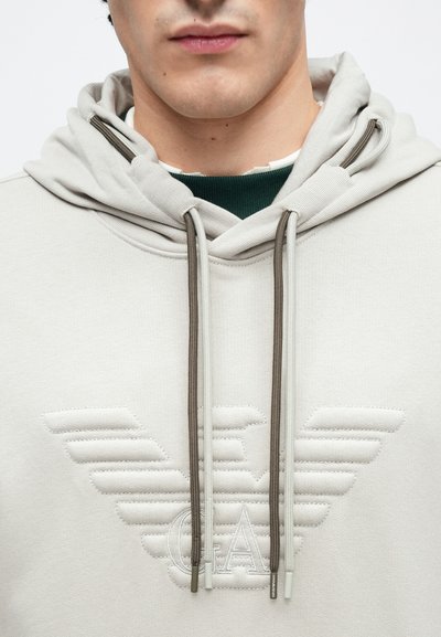 Φωτεινή γκρι φούτερ με κουκούλα, με ένα εξέχον κεντητό λογότυπο με σχέδιο φτερών. Ομαλή υφή, ρυθμιζόμενα κορδόνια σε μπεζ και γκρι χρώμα.