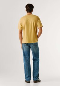 Camiseta de algodón amarilla con mangas cortas, combinada con unos jeans rectos de tiro medio en un tono azul claro que cuentan con un bolsillo trasero con parche y un leve desgaste.