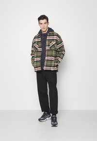 Casaco de flanela em padrão xadrez verde e vermelho, com dois bolsos no peito, sobre um hoodie escuro, combinado com calças pretas e ténis atléticos pretos.