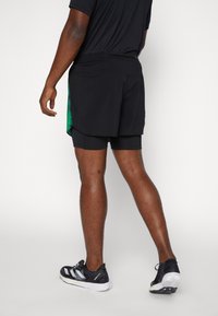 Svarta idrottsshorts med en åtsittande innerfoder, gröna accenter på sidan, elastisk midja och i kombination med svarta löparskor.