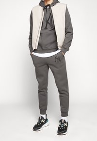 Sweat à capuche gris avec un grand logo noir, gilet beige sans manches et pantalon de survêtement gris assorti. Baskets noires avec semelles blanches et accents.