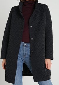 Sort quiltet jakke med høj krave, der har et tekstureret mønster og lynlås foran. Kombineret med en vinrød rullekrave og blå jeans.