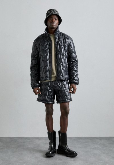 Schwarze Daunenjacke mit Zickzackmuster, kombiniert mit passenden Shorts. Dazu ein grünes Oberteil und schwarze Knöchelstiefel. Bucket Hat mit Karomuster.