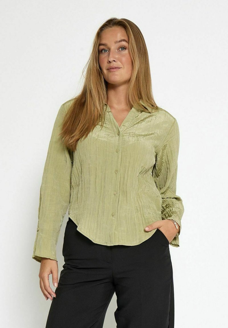 Donna con capelli lunghi biondi, che indossa una camicia button-up verde chiaro a trama e pantaloni neri, in posa con una mano in tasca su uno sfondo bianco.