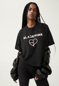 Černé tričko s krátkým rukávem s nápisem "BLACKPINK" v růžovém písmu a grafikou srdce, kombinované s černými skinny džíny a černou bomber bundou.