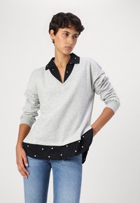 Maglione grigio con scollo a V e maniche lunghe. Colletto nero con pois bianchi, dotato di orlo a strati. Indossato con jeans blu.