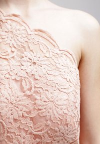 Lace top in lichtroze met bloemmotieven, één-schouder ontwerp, textuurstof en geschulpte randen. Close-up van het materiaal.