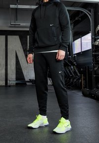 Czarna bluza Nike z fakturowanym materiałem i kieszenią kangurką, połączona z czarnymi dresami z białym logo. Jaskrawo żółte buty sportowe z niebieskimi akcentami.