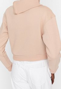 Sudadera corta de suave tela rosa con cuello alto y dobladillo acanalado, combinada con pantalones blancos que tienen bolsillos traseros.
