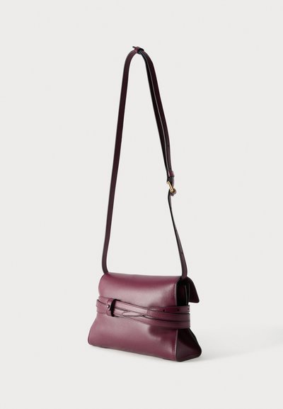 Μικρή σκούρα μοβ δερμάτινη τσάντα ώμου με ρυθμιζόμενο λουρί και διακοσμητική ζώνη μπροστά, εμφανίζεται κρεμασμένη πάνω σε ανοιχτό φόντο.