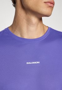 Nærbillede af en persons hals og underansigt iført en lilla tekstureret Salomon T-shirt med rund halsudskæring.