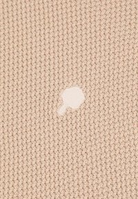 Beige gestrickter Stoff mit einem strukturierten Muster, das in der Mitte einen kleinen weißen unregelmäßigen Fleck oder ein Loch aufweist.