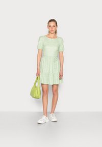 Vestido floral verde com mangas bufantes e saia em camadas, combinado com uma bolsa verde clara e ténis brancos. Tecido suave, design casual.