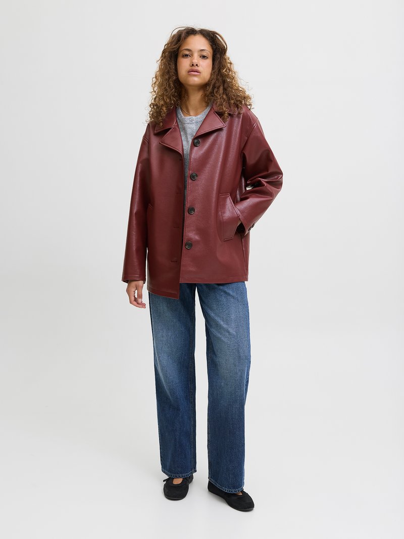 Chaqueta de imitación cuero burdeos con cuello, botones frontales y bolsillos laterales. Llevada sobre una blusa gris y combinada con vaqueros de pierna ancha azules.