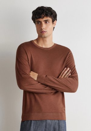 Jonge man met krullend donker haar, die een bruin shirt met lange mouwen en grijze broek draagt, staat met gekruiste armen en kijkt opzij.