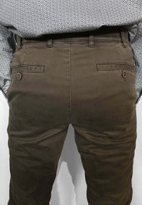 Pantaloni di cotone marrone con tasche posteriori dotate di chiusure con bottoni. Il tessuto ha una texture liscia e un design aderente.