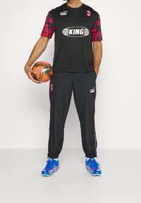 Svart fotbollskit med röda mönstrade ärmar, iförd blå skor. Håller i en orange och svart fotboll. Byxorna är svarta med röda accenter.