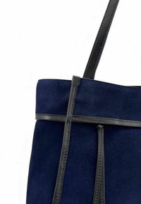 Sac fourre-tout en daim bleu marine avec accents en cuir noir, présentant un design plat, une lanière supérieure et deux pampilles suspendues pour plus de détails.