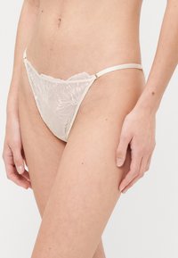 Lace brief in light beige, featuring floral embroidery, adjustable satin straps, and scalloped edges for a delicate design.