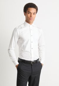 Fehér ing, hegyes gallérral, elülső gombolással fekete gombokkal, testhez álló kivitelben, sötét nadrággal és fekete övvel párosítva.