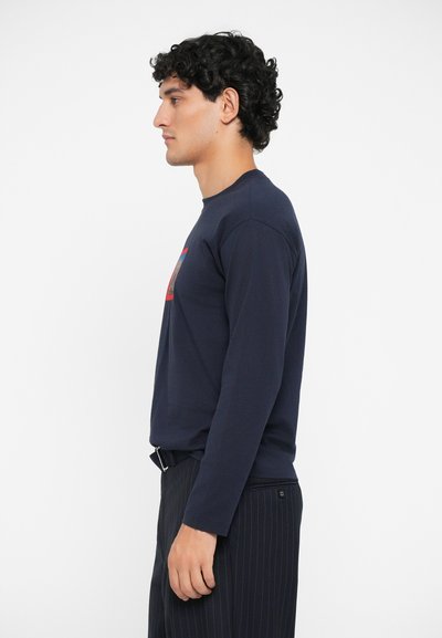 Navy long-sleeve T-shirt with round neckline, made of cotton. Features a graphic design in red and blue at the chest. Paired with pinstriped pants.