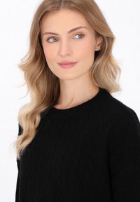 Zwarte kabelgebreide trui met een ronde hals. Zachte textuur, met een geribde onderkant. Model heeft golvend blond haar en een neutrale achtergrond.