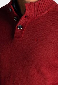 Rode gebreide trui met een geribbelde kraag, voorzien van een twee-knopen sluiting en subtiele logo borduursel aan de voorkant. Zachte textuur.
