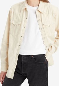 Beige painettu paita, jossa on länsimainen yoke-muotoilu, napilliset kiinnikkeet, kaksi rintataskua, käytetty valkoisen t-paidan päällä, yhdistetty tummiin farkkuihin.