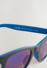 Occhiali da sole per bambini con montatura in plastica blu e accenti grigi. Le lenti hanno una sfumatura viola. Il testo inciso include "CE UK CA MADE IN CHINA 3-6YRS."