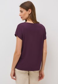 Purple short-sleeve top with a round neckline, smooth texture, and subtle shoulder detailing. Paired with light beige pants, shown from the back.