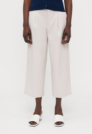 Pantalons larges beige clair avec une texture lisse et un devant plissé ; portés avec des sandales à talons blocs ouvertes blanches.