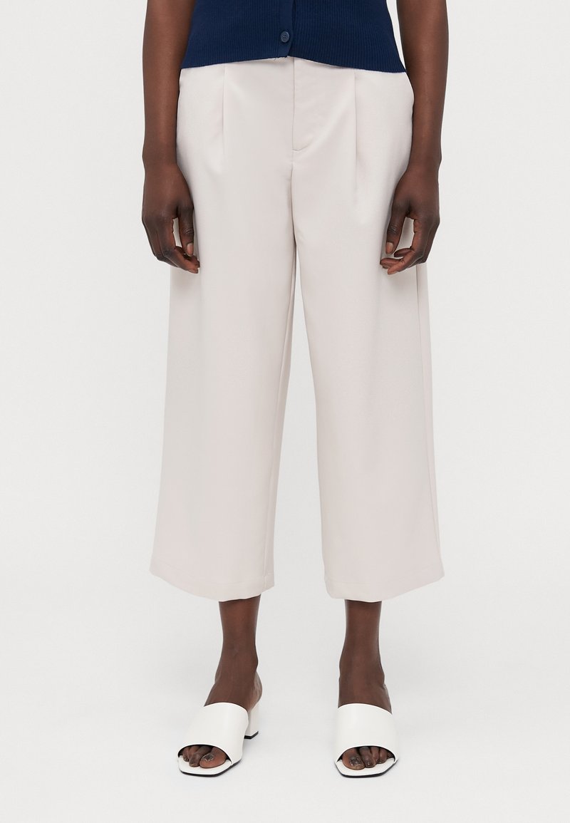 Lichtbeige wijde broek met een soepele textuur en geplooide voorkant; gedragen met witte open teen blokhak sandalen.