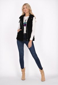 Vest din blană sintetică neagră cu închidere prin fermoar, asortat cu un top alb cu mânecă lungă și blugi skinny închisi la culoare. Botine tan cu design perforat.